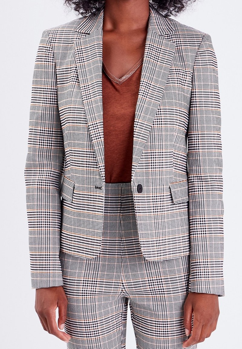 Femme portant un haut marron à col en V sous un blazer à carreaux gris et noir et pantalon taille haute assorti.