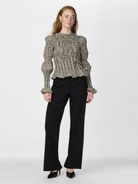 Žena stojaca, oblečená v kockovanej blúzke s dlhými rukávmi a volánikmi, čiernych nohaviciach s širokými nohavicami a čiernych lodičkách s špicatou špičkou na bielom pozadí.