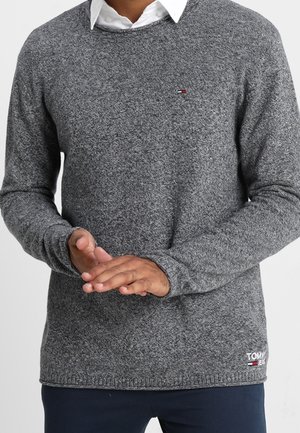 Grauer Strickpullover mit rundem Ausschnitt, gerippten Bündchen und Saum, mit einem kleinen Logo auf der Brust, über einem weißen Hemd getragen.