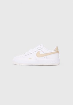 Weißer flacher Nike-Sneaker mit beigem Swoosh-Logo und Fersenlasche, mit weißen Schnürsenkeln und perforierter Zehenbox auf einfarbigem Hintergrund.