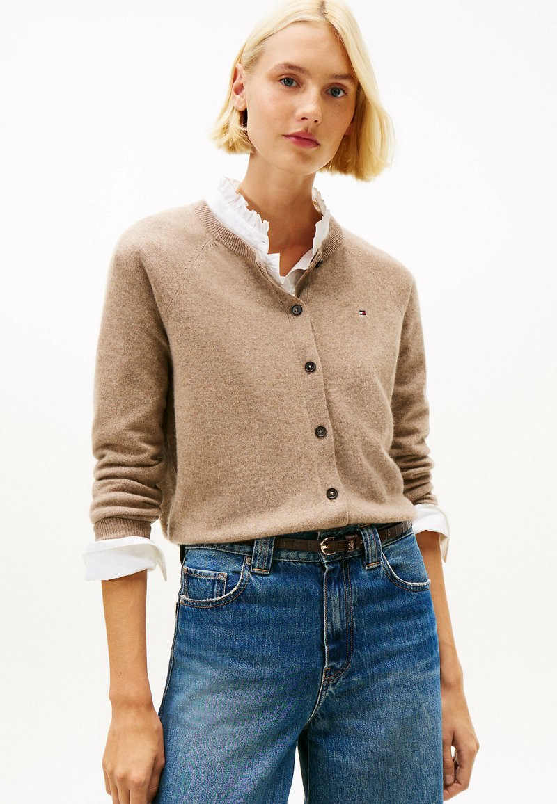 Jasnobrązowy kardigan z dekoltem w kształcie litery V i zapięciem na guziki, z prążkowanymi mankietami i małym haftowanym logo. Nosi się go na białej koszuli z falbanami.