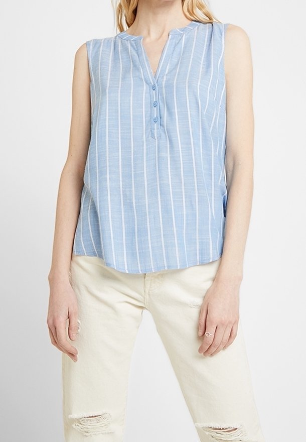Femme portant un chemisier sans manches bleu clair à rayures avec des boutons et un jean beige clair déchiré sur un fond uni.