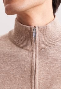 Nahaufnahme eines Halses, der einen beigen, gerippten Strick-Zipfel-Pullover mit einem teilweise geschlossenen silbernen Reißverschluss trägt.
