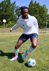 White hooded sweatshirt, navy shorts, and white socks with black stripes. Wearing bright blue and pink soccer cleats, dribbling a soccer ball on grass.