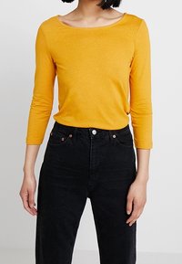 Žlutý top s dlouhými rukávy a širokým výstřihem, kombinovaný s černými džíny s vysokým pasem, které mají zapínání na knoflík a klasický pětikapsový design.