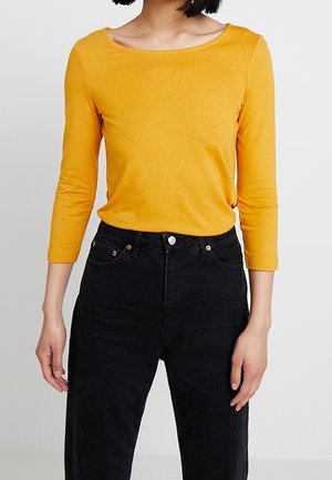 Haut jaune à manches longues avec un large décolleté, associé à un jean noir taille haute avec un devant à boutons et un design classique à cinq poches.