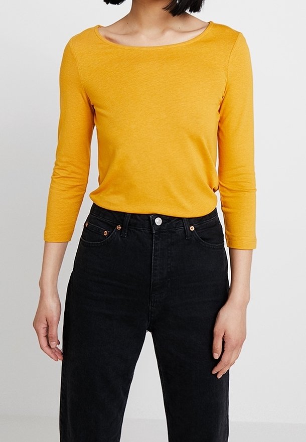 Žlutý top s dlouhými rukávy a širokým výstřihem, kombinovaný s černými džíny s vysokým pasem, které mají zapínání na knoflík a klasický pětikapsový design.