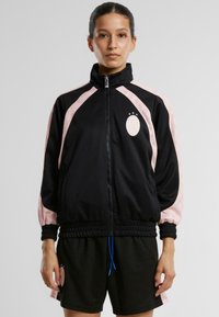 Črna zip-up jakna s svetlo roza vzorčastimi rokavi, elastičnimi manšetami in pasom. Značilna je okrogla nalepka na prsih s tremi zvezdami nad njo.