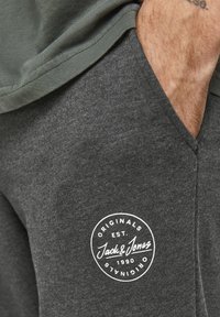 Graue Jogginghose mit strukturierter Oberfläche und einem kreisförmigen weißen Logo mit den Details "Jack & Jones" und "Established 1990" auf dem Oberschenkel.