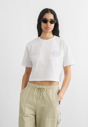 Νεαρή γυναίκα που φοράει γυαλιά ηλίου, λευκό κροσέ μπλουζάκι με μικρό σχέδιο και μπεζ παντελόνι με κορδόνι, με τα χέρια στις τσέπες, μπροστά σε λευκό φόντο.