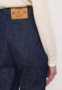 Mörkblå denimjeans med hjärtformad söm på bakfickorna. Läderpatch på midjan med logotyp. Långärmad krämfärgad topp synlig.