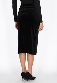Fekete bársony midi szoknya testhezálló sziluettel és finom fényességgel. Az alja egyenes, fekete, hegyes orrú magas sarkú cipővel párosítva.