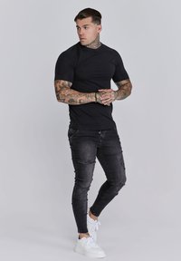 Fekete, rövid ujjú ing, skinny fekete farmernadrág, megviselt részletekkel, és fehér sportcipő. Az alakot tetoválások díszítik a karokon, semleges háttérrel.