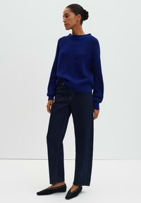 Pull en tricot bleu à manches larges, associé à un jean droit en denim foncé et des chaussures plates noires ornées de clous décoratifs.