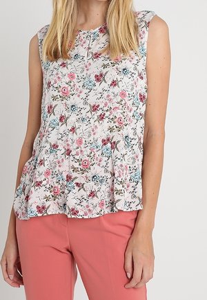 Blouse blanche sans manches avec imprimé floral rose, bleu et rouge, associée à un pantalon rose saumon porté par une femme debout.