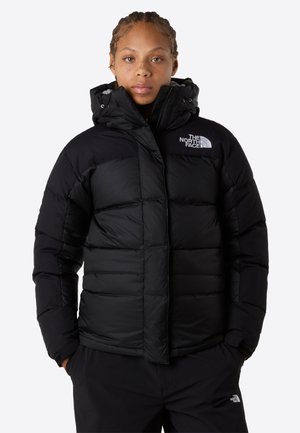 Persona vistiendo una chaqueta acolchada negra con capucha y el logo de The North Face, con las manos en los bolsillos, de pie frente a un fondo blanco.