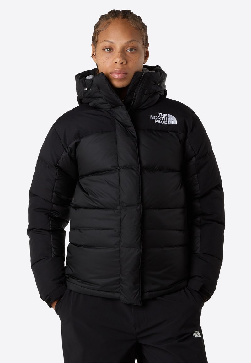 Persona vistiendo una chaqueta acolchada negra con capucha y el logo de The North Face, con las manos en los bolsillos, de pie frente a un fondo blanco.