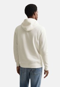 Camisola branca com capuz, ajuste casual, punhos e bainha canelados. Usada com jeans azul claro, mostrando uma vista traseira simples.