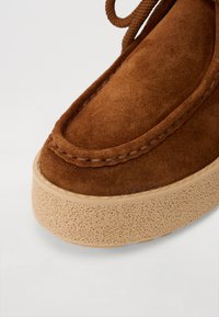 Chaussure en daim marron avec une pointe arrondie, des coutures décoratives et une semelle en caoutchouc épaisse et texturée de couleur crème. Les lacets sont noués en haut.