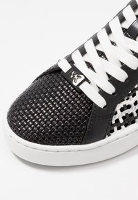 Baskets noires et blanches avec un embout en texture tissée, des lacets blancs et une étiquette métallique "MK" noire sur les lacets.