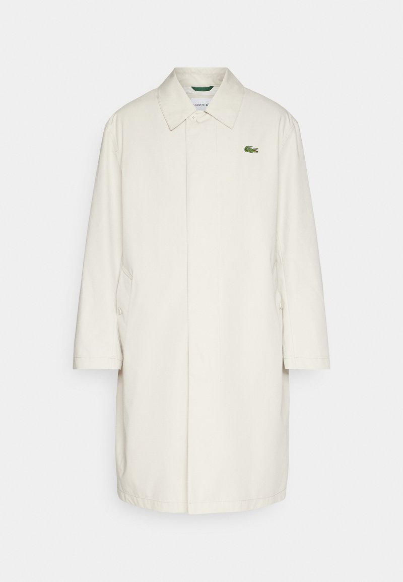 Impermeabile beige chiaro con colletto, maniche lunghe, tasche laterali e un accento di logo verde sul petto. Tessuto morbido al tatto.