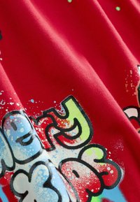 Rød bomuldsstof med farverigt graffiti-inspireret tekst og splattemønstre i blå, grøn og sort, der skaber et livligt design.