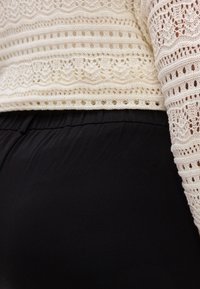 Πλεκτό πουλόβερ δαντέλα σε ελεφαντόδοντο με υφασμένους ανάγλυφους σχεδιασμούς, συνδυασμένο με μαύρο, ελαστικό παντελόνι. Το πουλόβερ έχει κυματοειδή σχεδίαση και ανοιχτές λεπτομέρει ες.