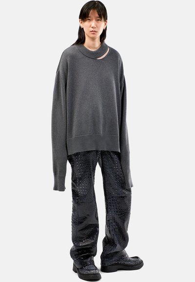 Grijze oversized trui met een ronde hals, gecombineerd met glanzende zwarte broek met een krokodillenpatroon. Chunky zwarte schoenen maken de look compleet.