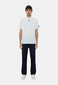 Polo bianco con colletto a zip, maniche corte e patch con logo; abbinato a jeans scuri e sneaker bianche. Cappellino nero indossato in avanti.