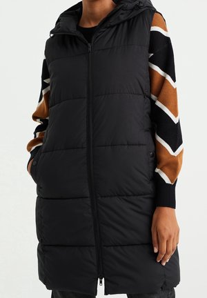 Persoon draagt een zwart lang doorgestikt vest met capuchon over een bruine, zwarte en witte zigzag-trui.