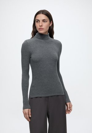 Femme aux longs cheveux foncés portant un pull à col roulé gris ajusté et un pantalon large foncé, debout devant un fond uni.