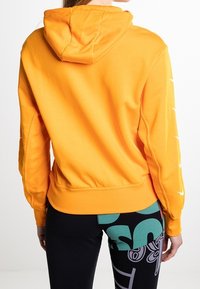 Oranžová mikina s kapucí a šňůrkou, žeberkovými manžetami a hladkým bavlněným směsným materiálem. Spárována s černými legínami s vzorem a zelenými akcenty.