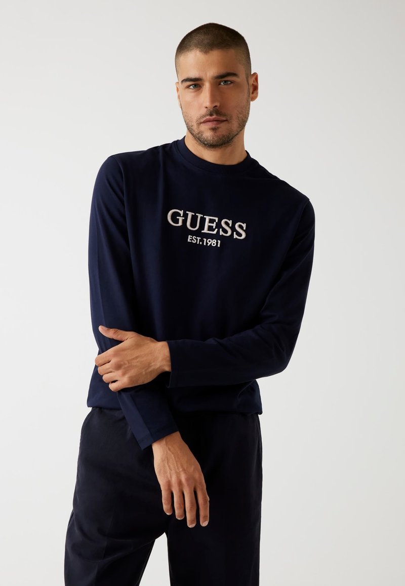 Camicia a maniche lunghe navy con "GUESS EST.1981" ricamato in beige sul petto. Materiale misto cotone con un design classico a girocollo.
