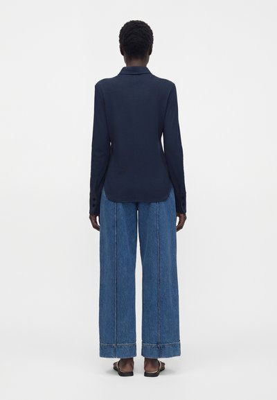 Personne portant un blazer bleu marine foncé et un jean bleu à jambes larges, debout, de dos, sur un fond blanc uni.