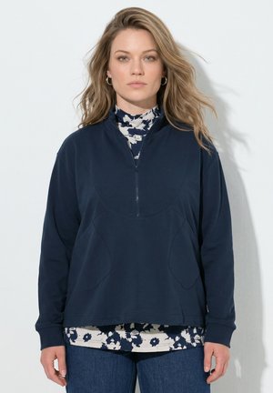 Kvinde med løst blondt hår iført en mørkeblå kvart-lyn pullover over en hvid og mørkeblå blomstret skjorte og blå jeans, stående mod en hvid væg.