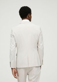 Chaqueta de traje de color beige claro, corte ajustado, diseño de un solo botón, solapas con muesca, dos aberturas traseras, detalles en botones dorados, estructura entallada.