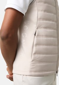 Beige gepolsterte Weste mit gestepptem Design, seitlichem Reißverschluss und eingeprägtem Logo. Weiches Material, nahtloses Design.