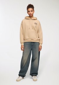 Felpa beige con tasca a marsupio e testo nero, abbinata a jeans in denim svasati sbiaditi e sneakers crema. Tessuto morbido, vestibilità rilassata.