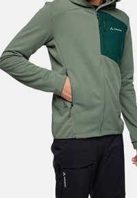 Felpa verde con zip, realizzata in tessuto texturizzato, con un pannello frontale in un verde scuro a contrasto e cappuccio regolabile con cordini. Tasche laterali in vista.