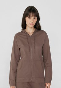 Sudadera con cremallera marrón, con capucha, que incluye bolsillos frontales, una textura suave y cordones. Diseño simple y moderno con mangas largas.