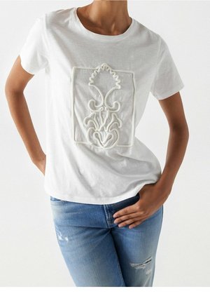 Person trägt ein weißes T-Shirt mit kurzen Ärmeln und erhabener Blumenstickerei in einem rechteckigen Rahmen, kombiniert mit hellblauen, zerrissenen Jeans.