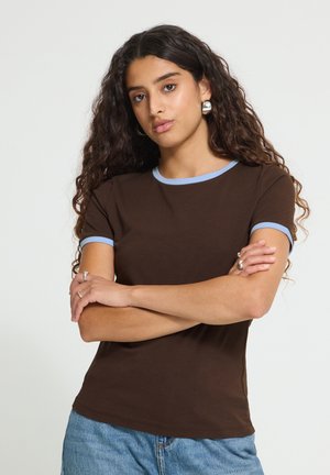 Jeune femme aux longs cheveux bouclés portant un t-shirt marron avec des bordures bleu clair et un jean bleu, debout les bras croisés.