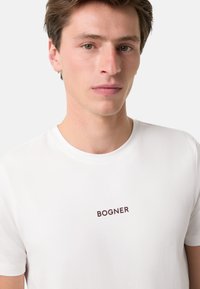Bílý bavlněný tričko s kulatým výstřihem a krátkými rukávy, na hrudi s vyšitým nápisem "BOGNER" v tmavě hnědé barvě.
