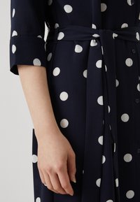 Rochie bleumarin cu buline albe, dintr-un material texturat, cu închidere prin buton și curea din același material. Mâneci cu manșetă adaugă detalii designului.