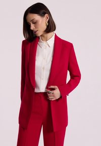 Jasnoczerwony żakiet o dopasowanym kroju z kołnierzem w stylu szala, zestawiony z białą koszulą na guziki. Posiada eleganckie linie i dopasowaną sylwetkę, bez widocznych wzorów.
