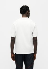 Άσπρο βαμβακερό μπλουζάκι με κοντά μανίκια, στρογγυλή λαιμόκοψη και απλό σχέδιο, που φοριέται από έναν άνθρωπο που είναι γυρισμένος με την πλάτη, σε συνδυασμό με μαύρο παντελόνι με κόκκινες λεπτομέρειες.