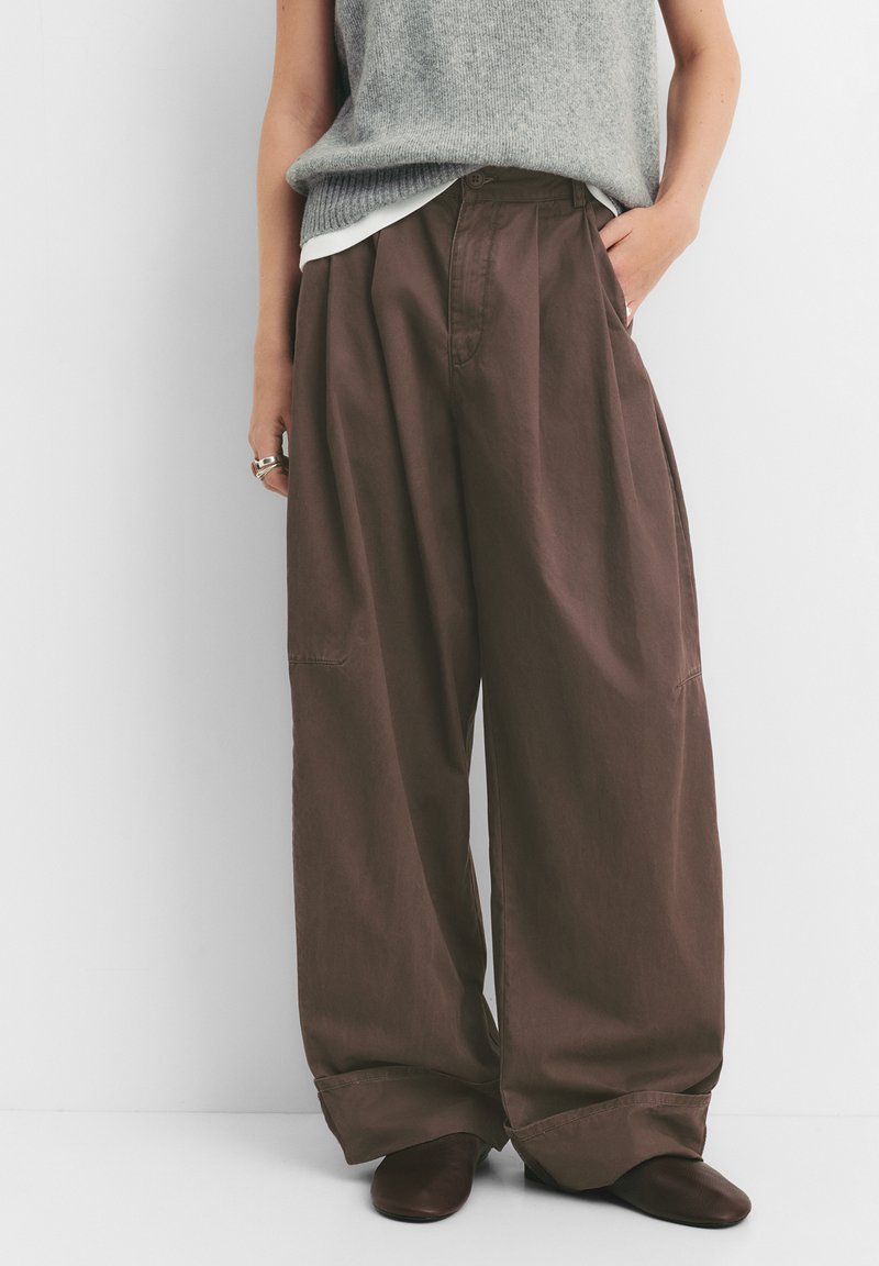 Bruine wijde pantalons van lichtgewicht stof met voorplooien, zijzakken en een omgeslagen zoom. Draagt een grijze mouwloze top.
