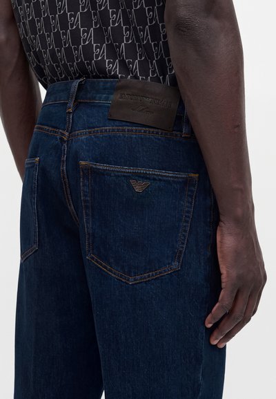 Σκούρο μπλε τζιν με καφέ ραφές και λογότυπο Emporio Armani στην πίσω τσέπη και δερμάτινη ετικέτα μάρκας στη ζώνη, φορεμένα από ένα άτομο.