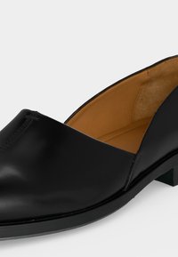 Μαύρο δερμάτινο παπούτσι τύπου slip-on με χαμηλό τακούνι και ταμπά εσωτερική επένδυση, εμφανίζεται από εμπρόσθια πλευρική γωνία σε λευκό φόντο.