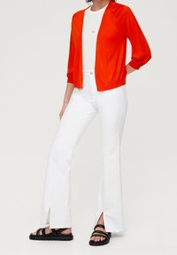 Czerwony kardigan na białej koszuli, zestawiony z białymi dzwonowatymi dżinsami z bocznymi rozcięciami oraz czarnymi sandałami na paski. Gładkie tekstury, casualowy design.